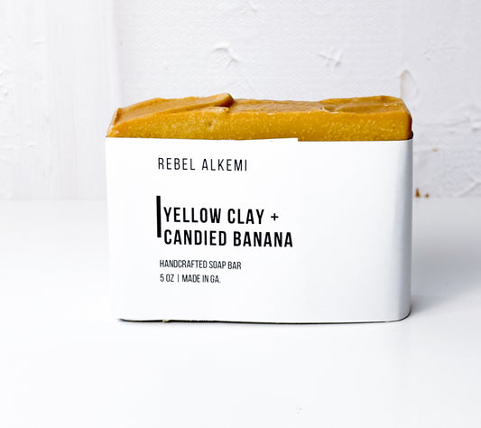 Handcrafted soap bar with 'Yellow Clay + Candied Banana' scent by Rebel Alkemi on a white background.